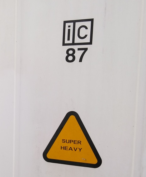 Zdjęcie przedstawia naklejki na ścianie kontenera, jedna o napisie „IC 87” a druga „Super Heavy” w żółtym trójkącie